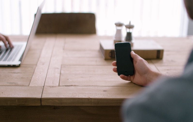 Man holding phone sitting next to woman on computer at wooden desk